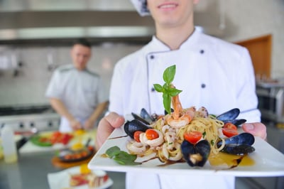 Handsome chef dressed in white uniform decorating pasta salad and seafood fish in modern kitchen