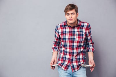 Poor handsome young man in checkered shirt and jeans showing empty pockets over grey background